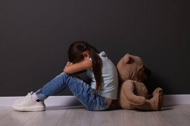 Child abuse. Upset little girl with teddy bear sitting on floor near gray wall indoors