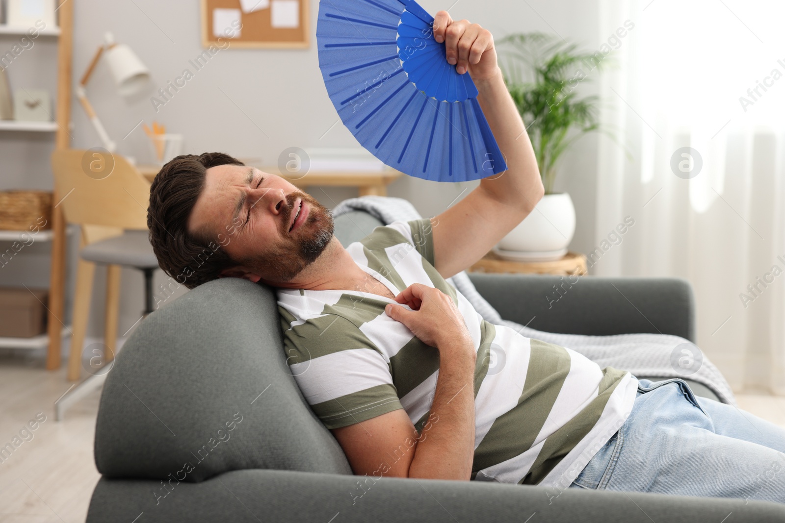 Photo of Bearded man waving blue hand fan to cool himself on sofa at home