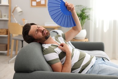 Photo of Bearded man waving blue hand fan to cool himself on sofa at home