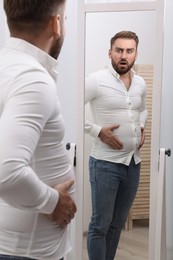 Photo of Scared man wearing tight shirt in front of mirror at home. Overweight problem