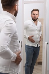 Scared man wearing tight shirt in front of mirror at home. Overweight problem