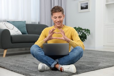 Photo of Happy young man having video chat via laptop and making heart with hands on carpet indoors. Long-distance relationship