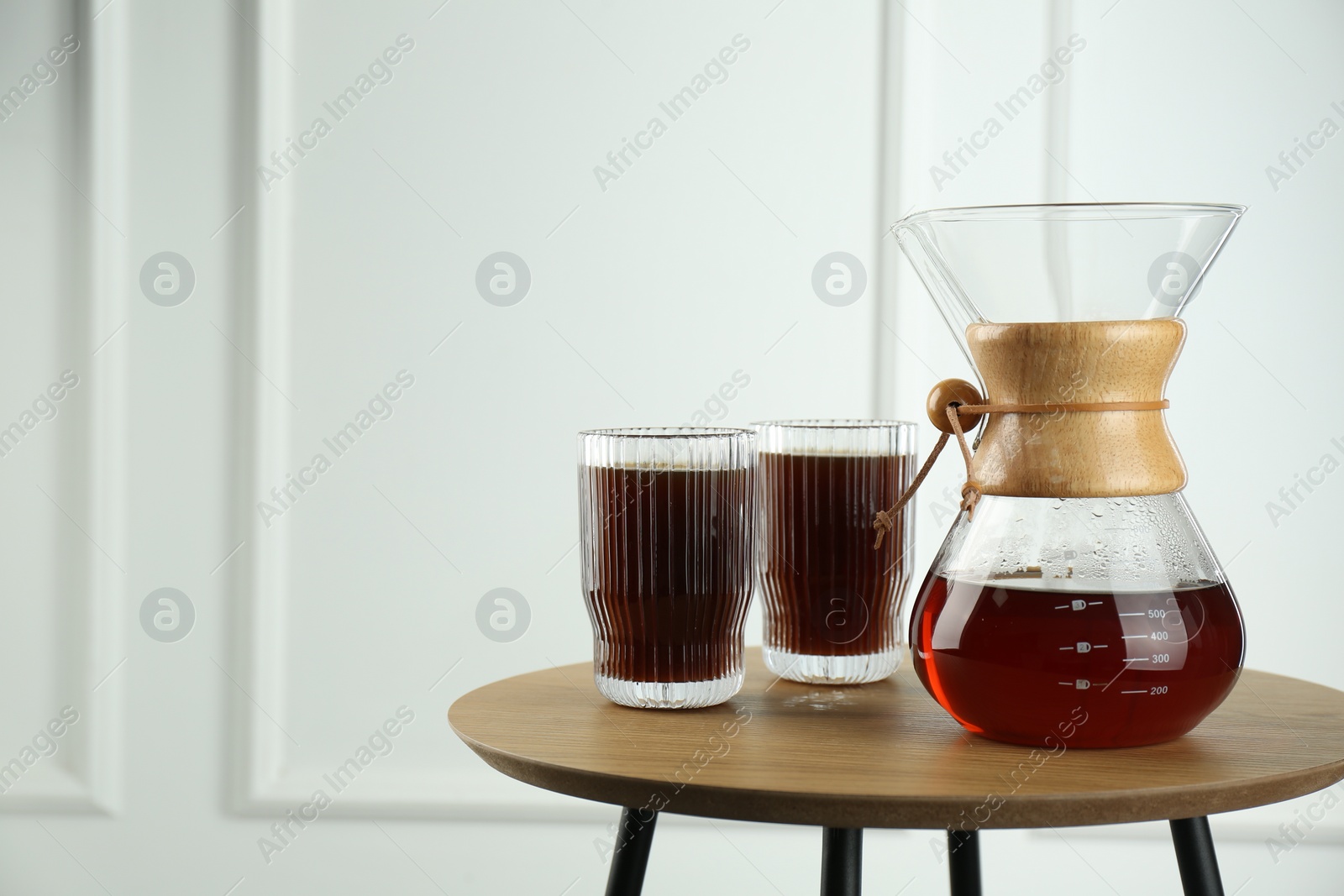 Photo of Glass chemex coffeemaker and glasses of coffee on wooden table against white wall, space for text