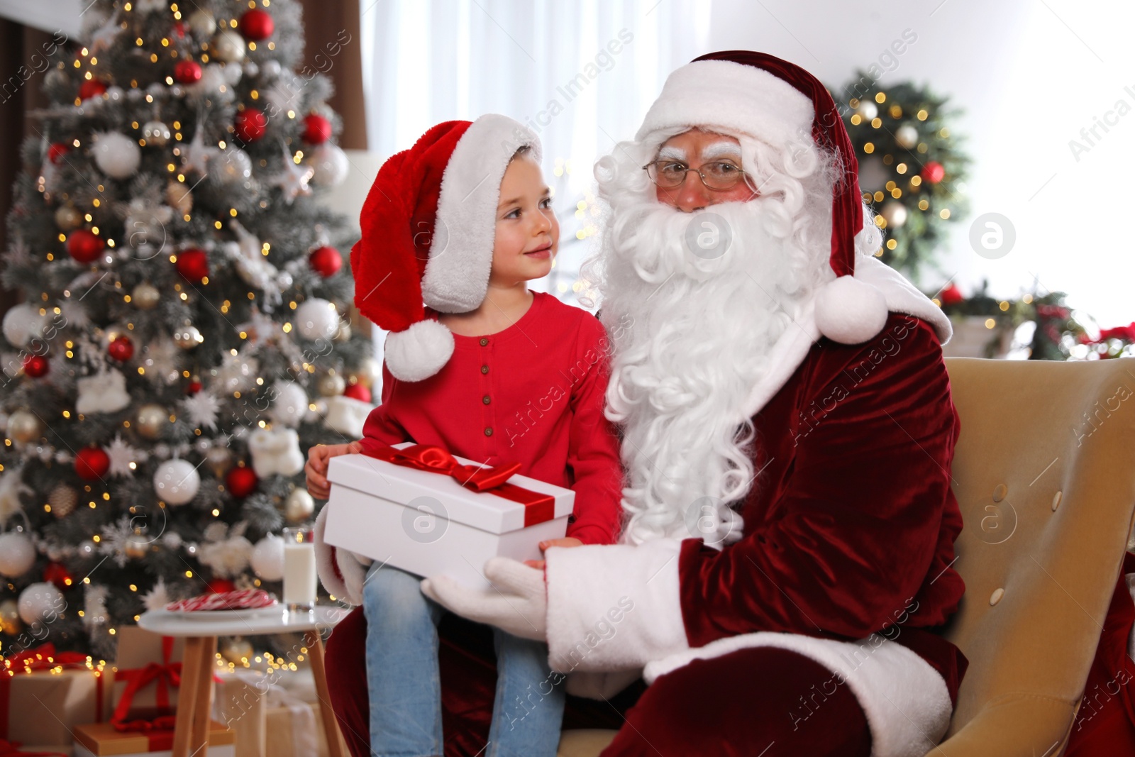 Photo of Santa Claus giving present to little girl in room decorated for Christmas