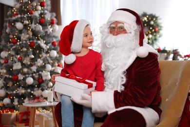 Santa Claus giving present to little girl in room decorated for Christmas