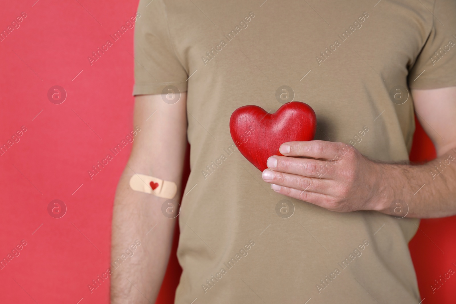 Photo of Blood donation concept. Man with adhesive plaster on arm holding red heart against color background, closeup