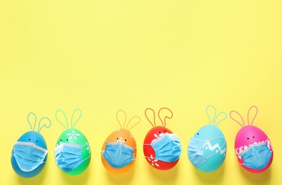 Image of COVID-19 pandemic. Colorful Easter eggs with cute bunny ears in protective masks and space for text on yellow background, flat lay