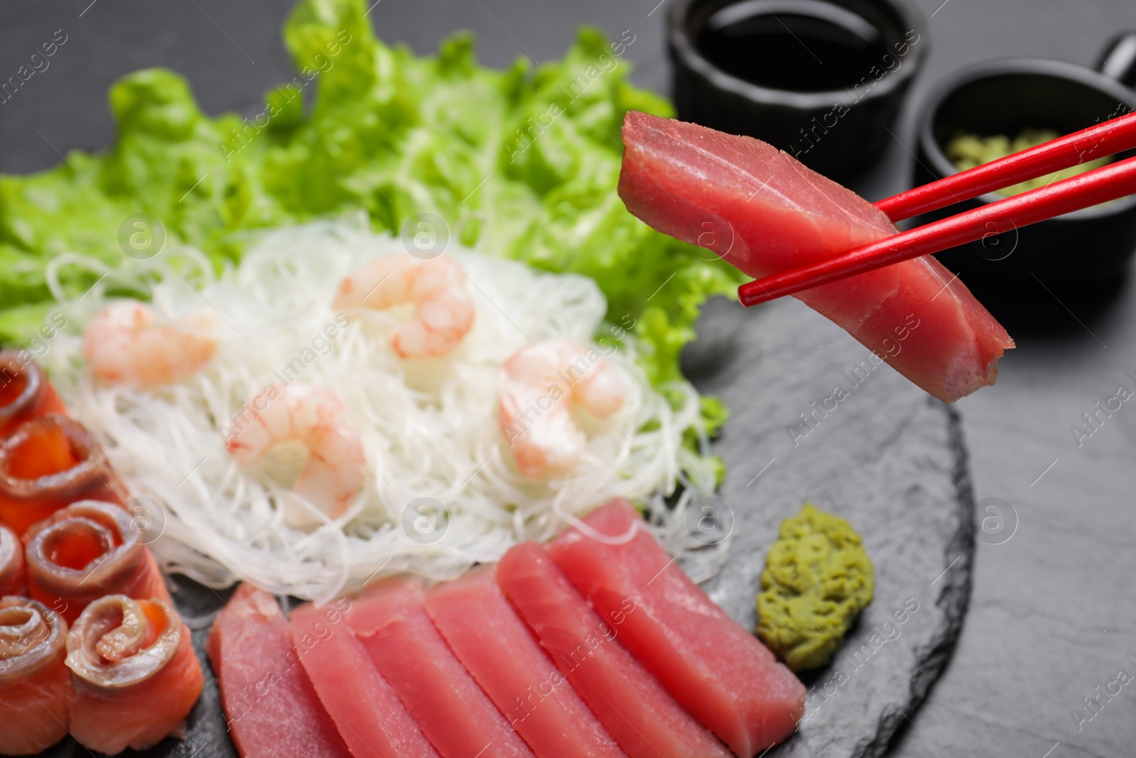 Photo of Sashimi set (raw tuna, salmon slices and shrimps) served with funchosa, lettuce and vasabi on dark table, closeup. Chopsticks with fish