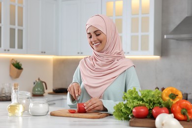 Muslim woman making delicious salad with vegetables at white table in kitchen