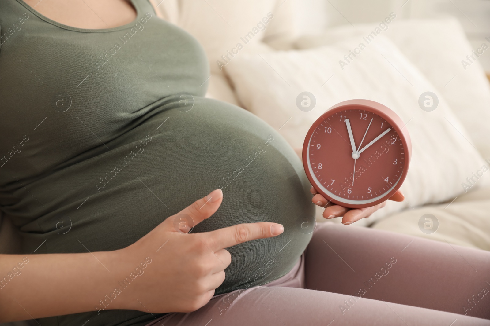 Photo of Young pregnant woman pointing at clock near her belly indoors, closeup. Time to give birth