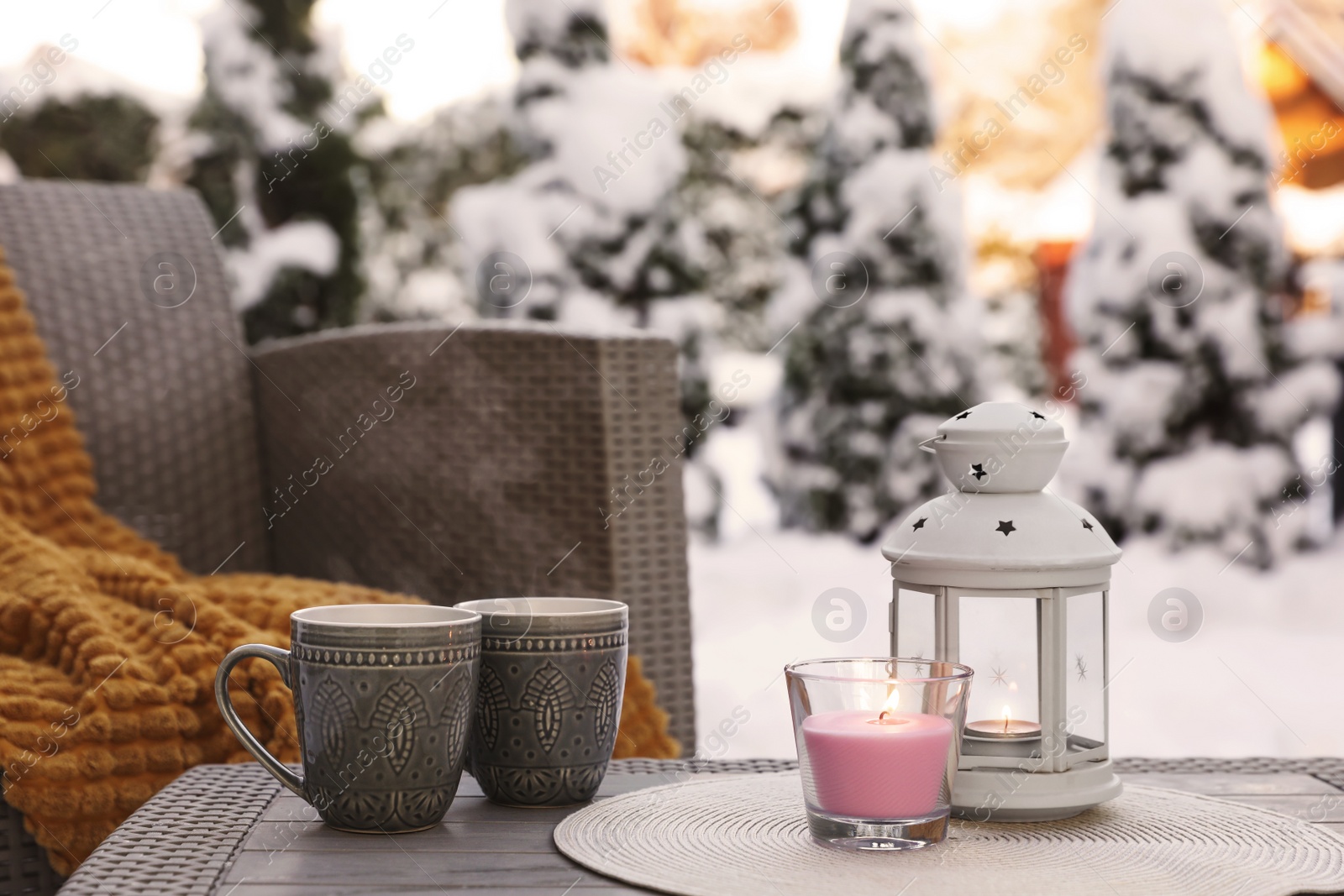 Photo of Burning candle, lantern and cups with hot drink on coffee table outdoors. Cosy winter