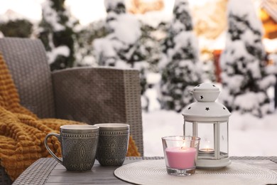 Photo of Burning candle, lantern and cups with hot drink on coffee table outdoors. Cosy winter