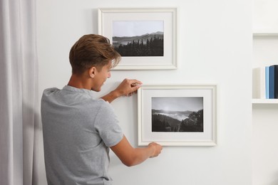Young man hanging picture frames on white wall indoors, back view
