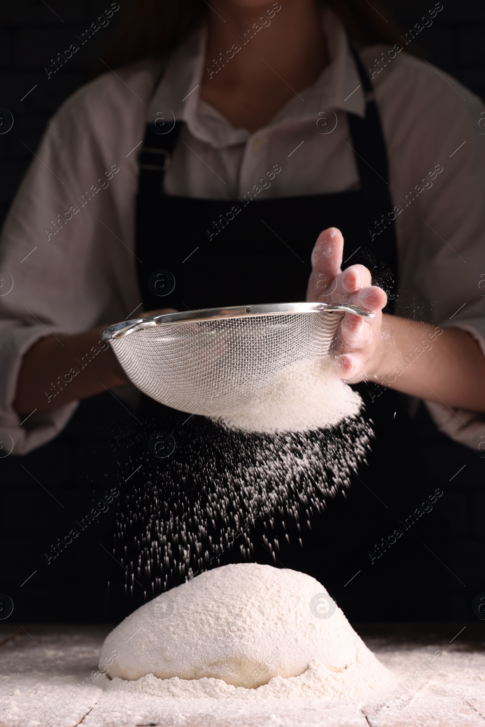 Photo of Woman sprinkling flour over dough at wooden table on dark background, closeup