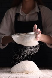 Woman sprinkling flour over dough at wooden table on dark background, closeup