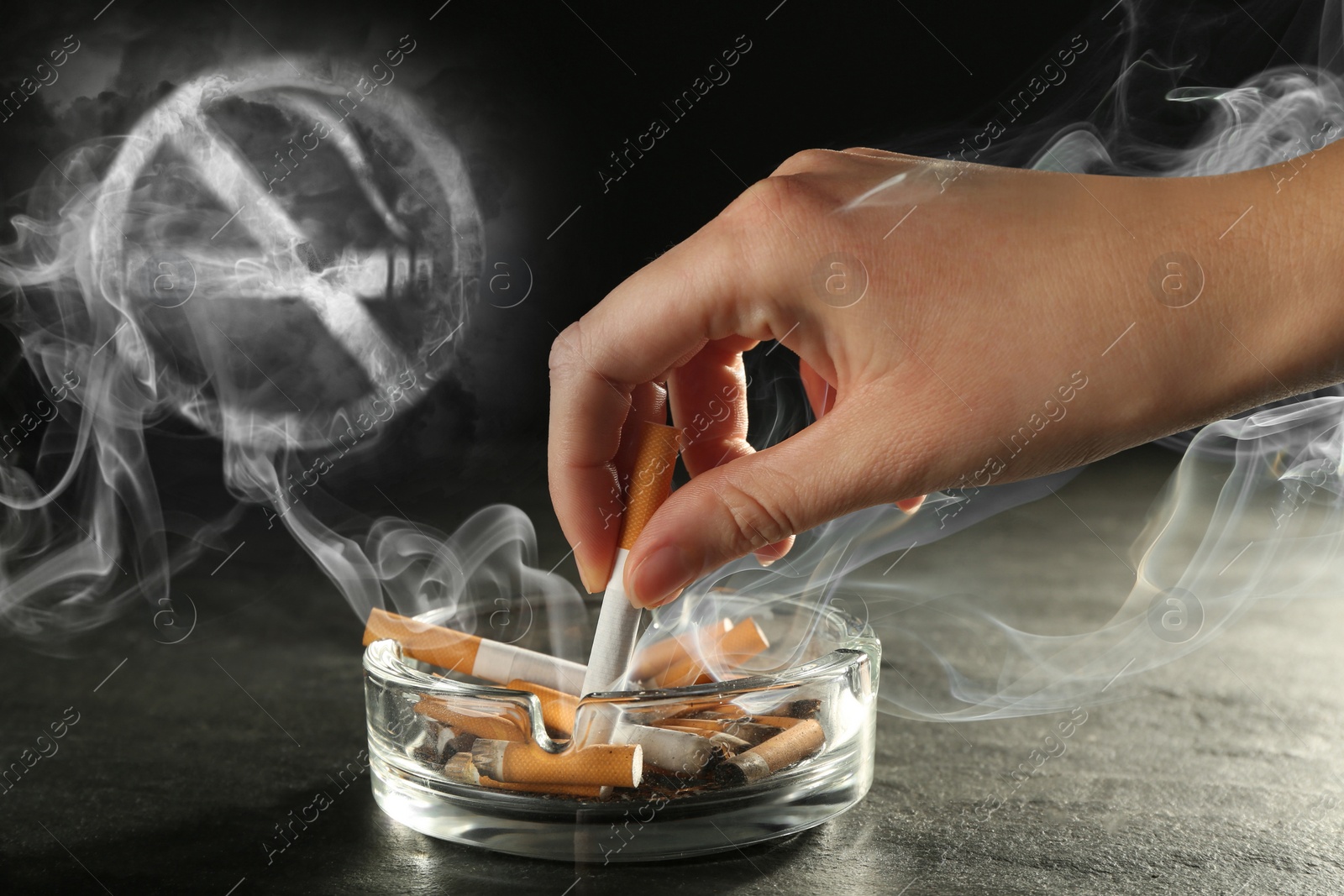 Image of No Smoking, round sign with crossed cigarette of smoke. Woman extinguishing cigarette in glass ashtray
at grey table against black background, closeup