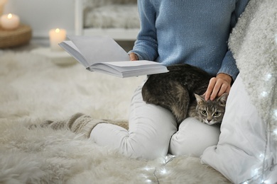 Photo of Young woman with cute cat reading book at home, closeup. Cozy winter