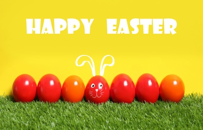 One egg with drawn face and ears as Easter bunny among others on green grass against yellow background