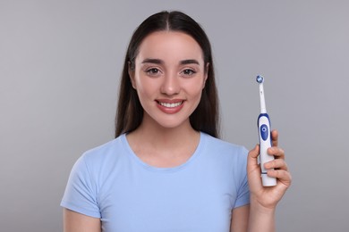 Happy young woman holding electric toothbrush on light grey background