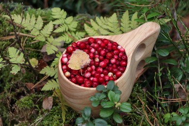 Photo of Many tasty ripe lingonberries and leaf in wooden cup near fern outdoors, closeup