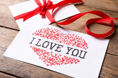 Card with text I Love You, envelope and red ribbon on wooden background, closeup