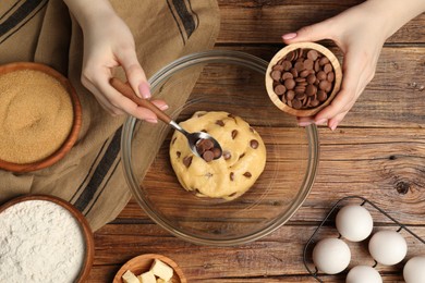 Photo of Cooking sweet cookies. Woman adding chocolate chips to dough at wooden table, top view