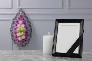Photo of Photo frame with black ribbon, burning candle on light grey table and wreath of plastic flowers near wall indoors, space for text. Funeral attributes