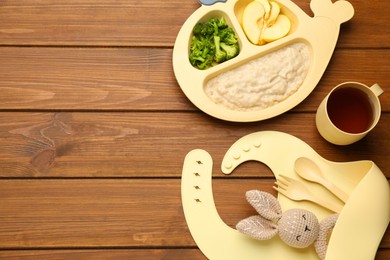 Photo of Silicone baby bib, toy and plastic dishware with healthy food on wooden table, flat lay. Space for text