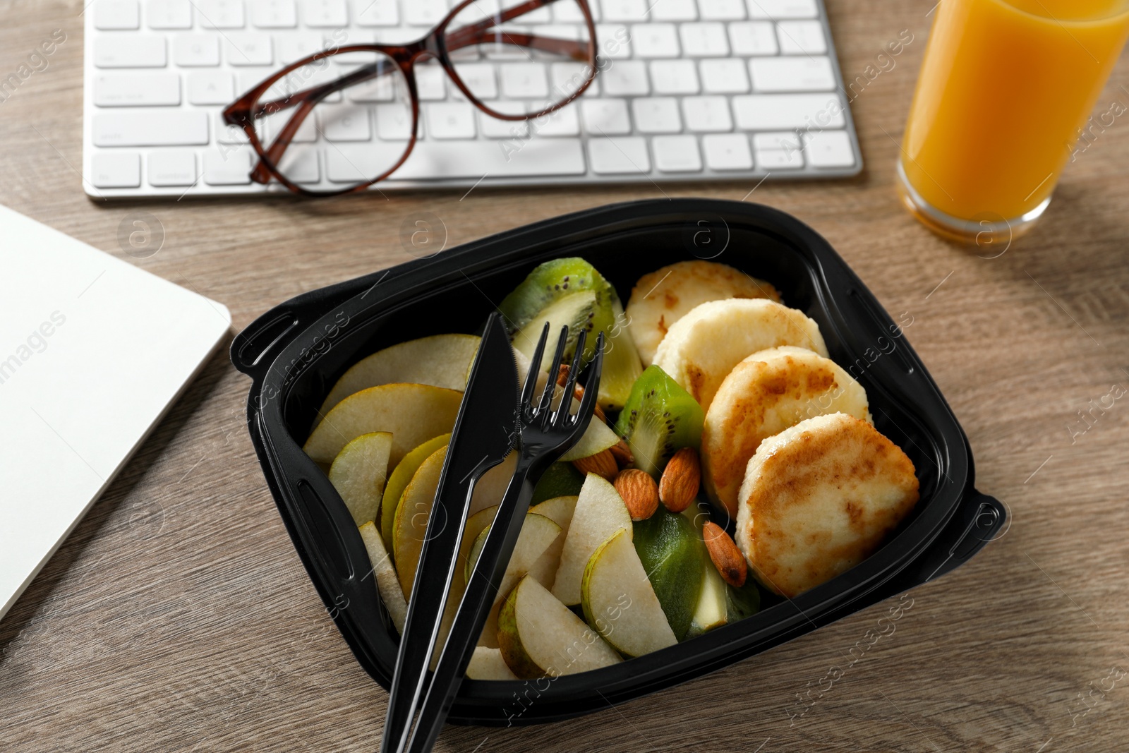 Photo of Container with tasty food, glass of juice, keyboard, cutlery and glasses on wooden table. Business lunch