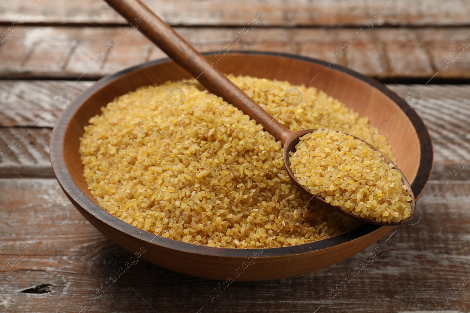 Photo of Bowl and spoon with raw bulgur on wooden table, closeup