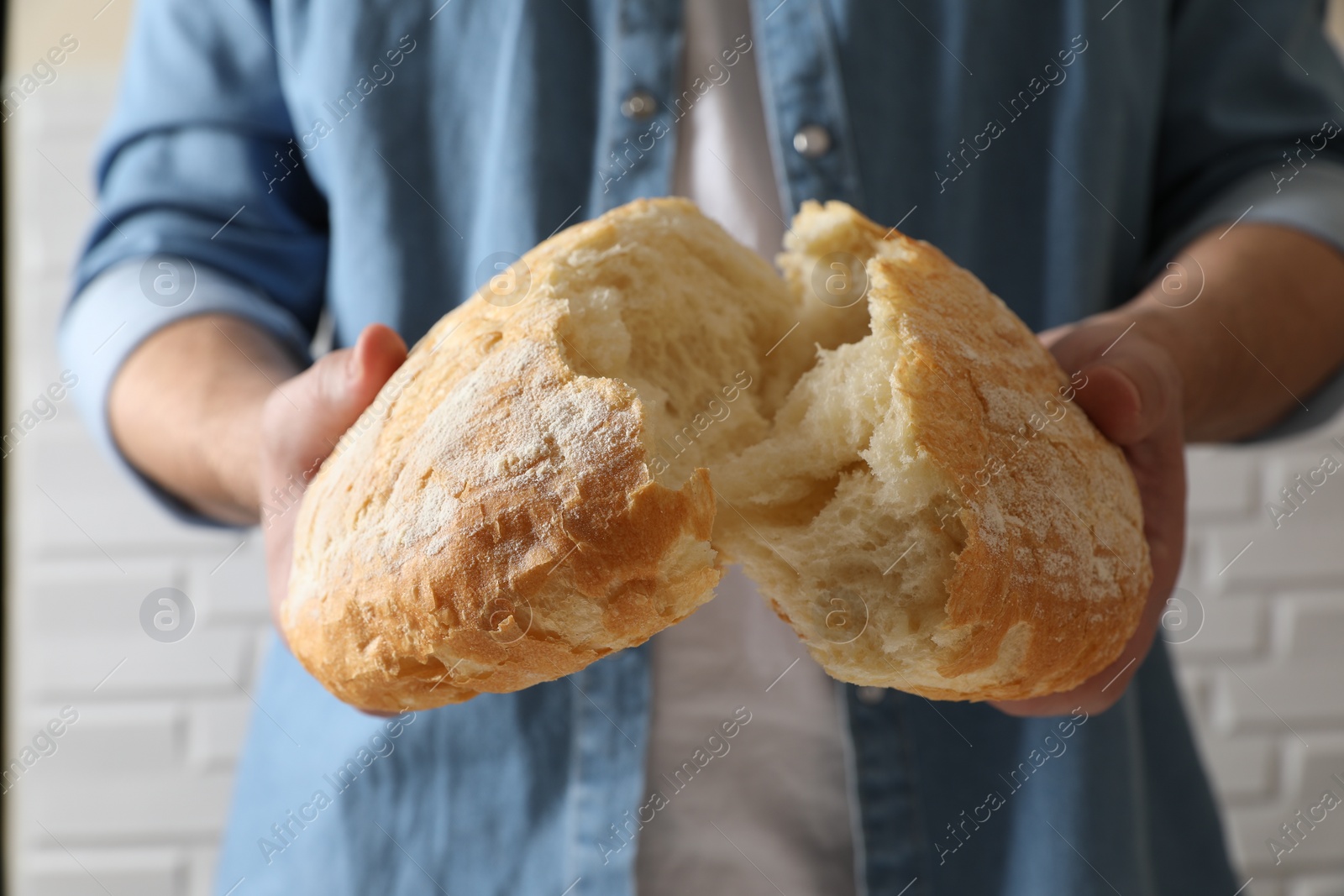 Photo of Man breaking loaf of fresh bread near white brick wall, closeup