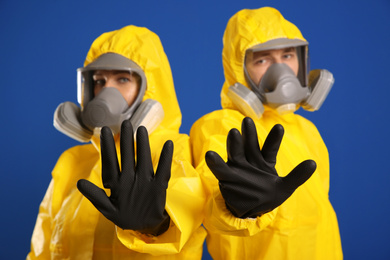 Photo of Man and woman in chemical protective suits making stop gesture against blue background, focus on hands. Virus research