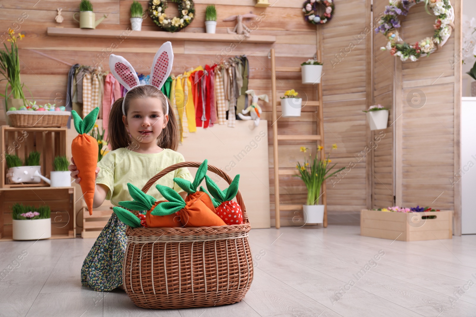 Photo of Adorable little girl with bunny ears and basket full of toy carrots in Easter photo zone