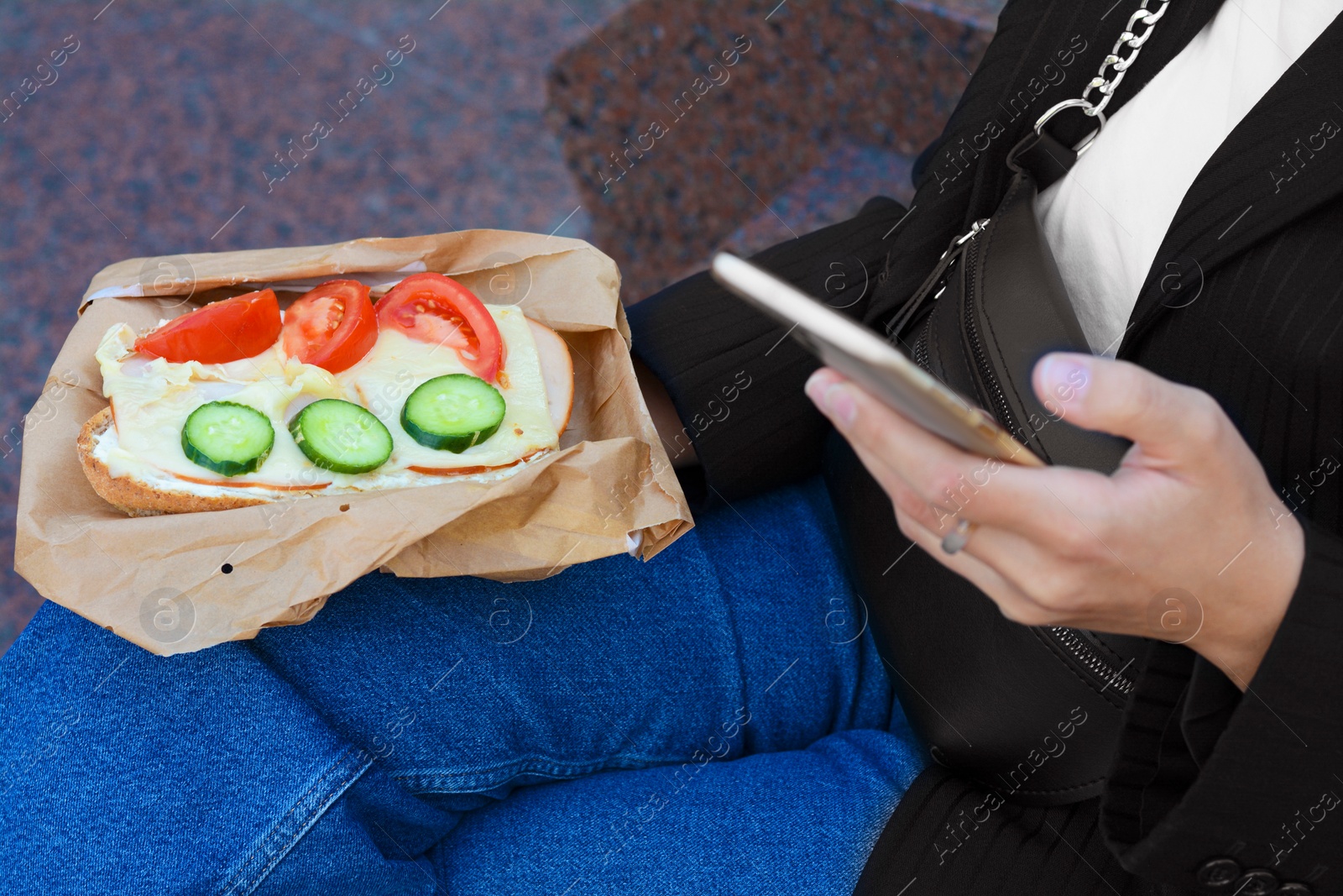 Photo of Woman holding tasty sandwich with vegetables and using phone outdoors, closeup. Street food