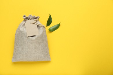 Photo of Tied burlap bag with tag and green leaves on yellow background, flat lay. Space for text