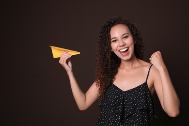 Beautiful African-American woman playing with paper plane on brown background