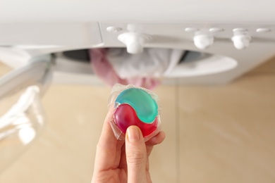 Photo of Woman holding laundry detergent capsule near washing machine indoors, top view