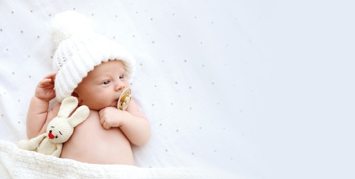 Image of Cute newborn baby in white knitted hat with toy lying on bed, top view with space for text. Banner design
