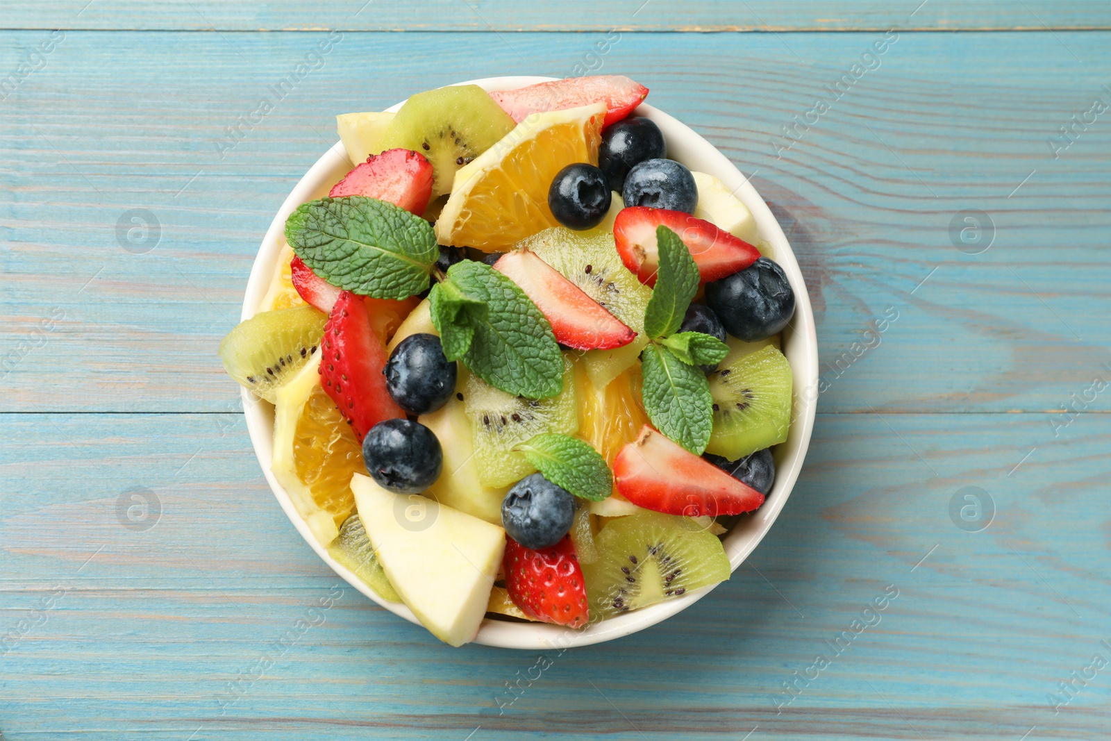 Photo of Tasty fruit salad in bowl on light blue wooden table, top view