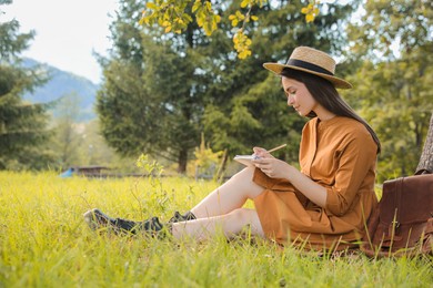 Photo of Beautiful young woman drawing with pencil in notepad outdoors on green grass