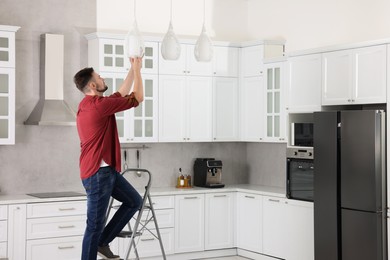 Photo of Young man installing ceiling lamp on stepladder in kitchen, space for text