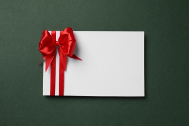 Photo of Blank gift card with red bow on dark green background, top view. Space for text