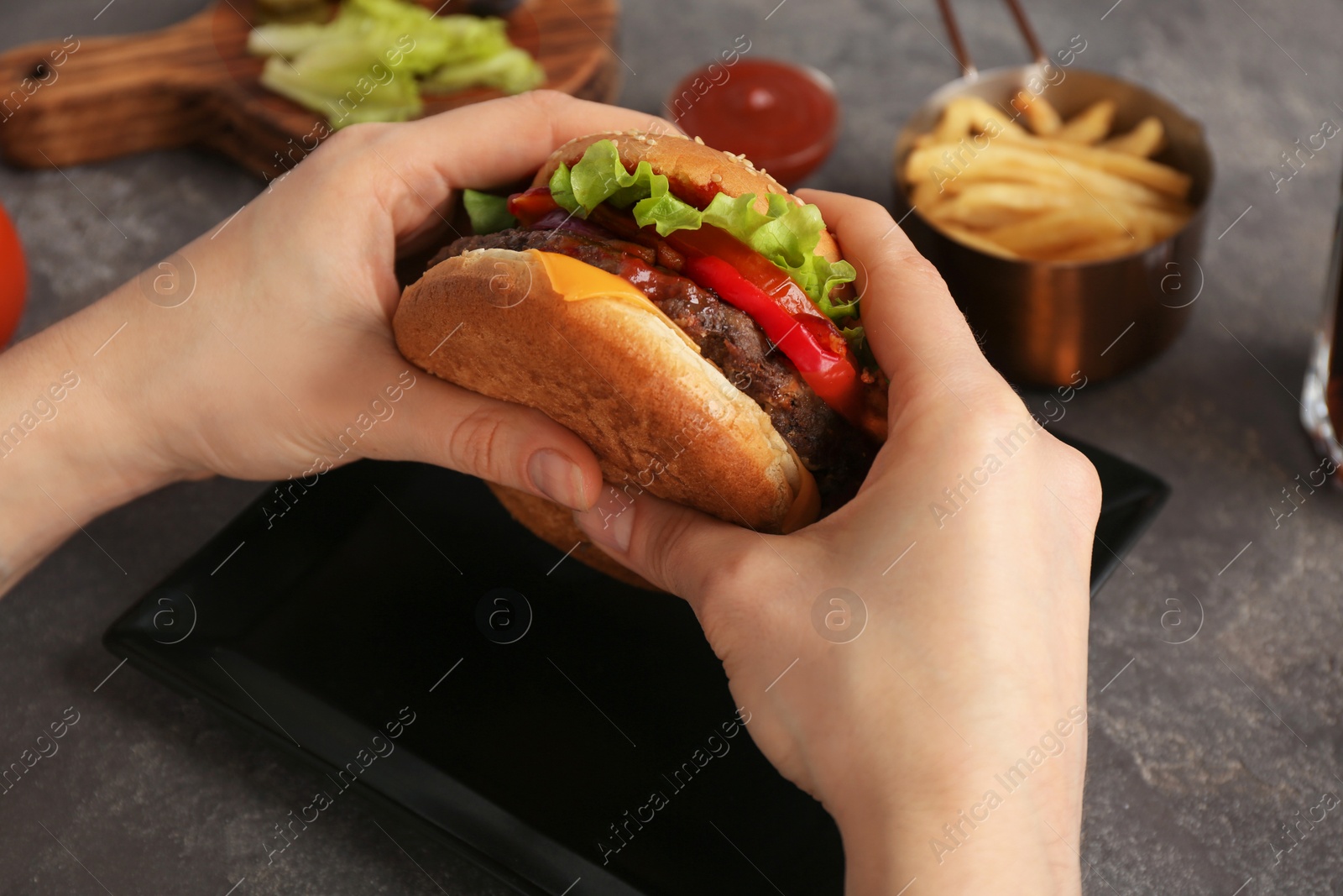 Photo of Woman holding tasty burger with bacon over grey table, closeup