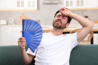 Bearded man waving blue hand fan to cool himself on sofa at home