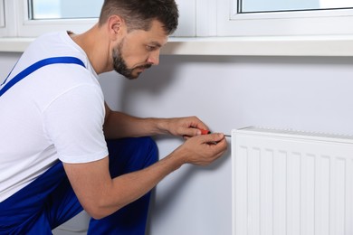 Photo of Professional plumber using screwdriver while preparing heating radiator for winter season indoors