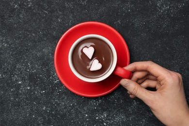 Photo of Woman holding cuphot chocolate with heart shaped marshmallows at grey table, top view