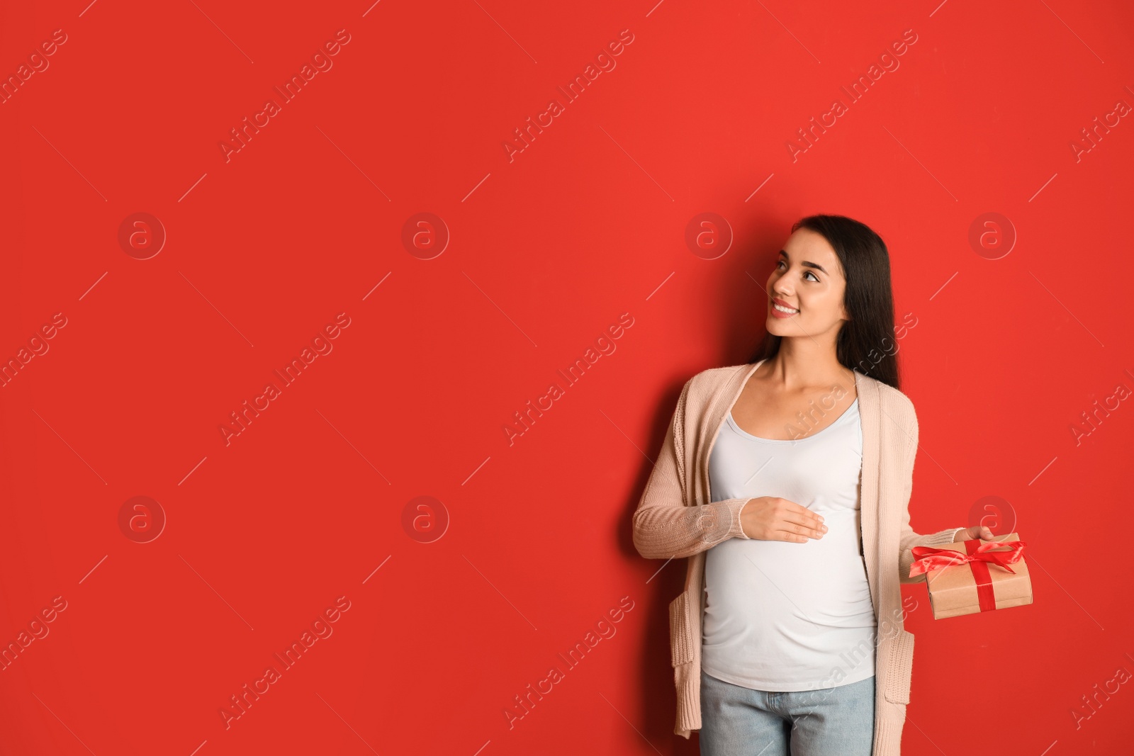 Photo of Happy pregnant woman with Christmas gift box on red background, space for text. Expecting baby