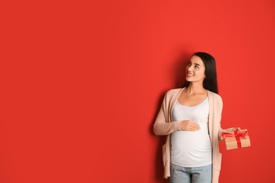 Happy pregnant woman with Christmas gift box on red background, space for text. Expecting baby