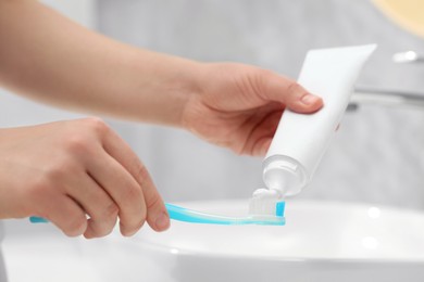 Photo of Woman squeezing toothpaste from tube onto toothbrush near sink in bathroom, closeup