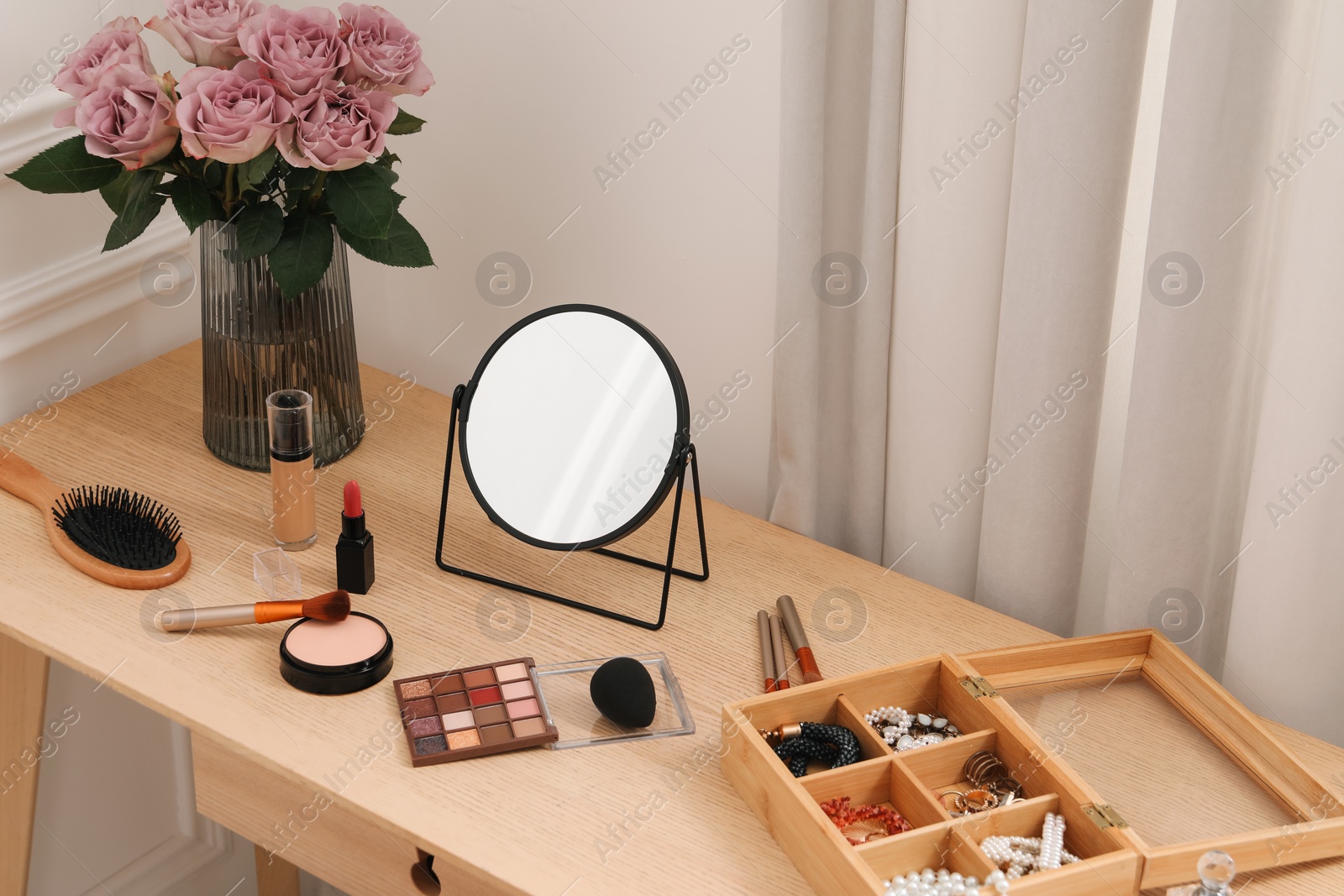 Photo of Mirror, cosmetic products, box of jewelry and vase with pink roses on wooden dressing table in makeup room